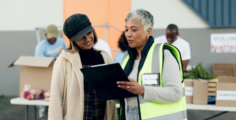 An older woman in a yellow vest and with a volunteer tag showing a tablet to a younger woman while standing outside with a bunch of boxes in the background.