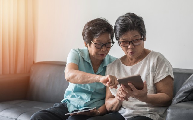 Two older women sitting on a grey couch and looking at a tablet.