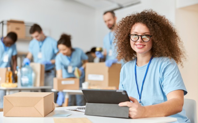 A young woman to the right staring at the camera and smiling while holding a tablet with people in the background going through boxes.